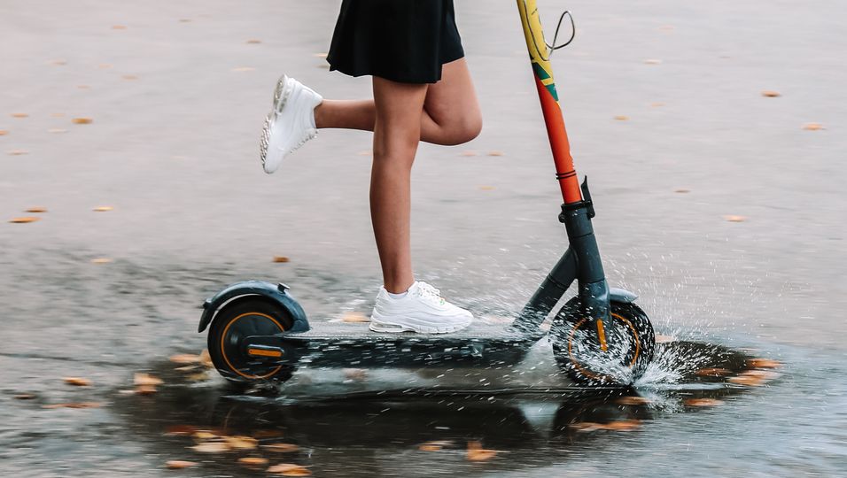 Frau fährt mit weißen Schuhen auf einem E-Scooter durch eine Pfütze