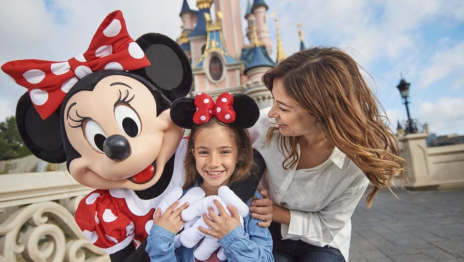 Das Bild zeigt ein Mädchen, das von Minnie und ihrer Mutter umarmt wird und vor dem Schloss in Disneyland® Paris posiert.