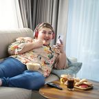 Übergewichtige Frau sitzt auf Sofa, schaut ins Smartphone und isst Chips.