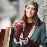 Eine junge Frau mit Einkaufstüten und Handy in der Hand lächelt in die Kamera.