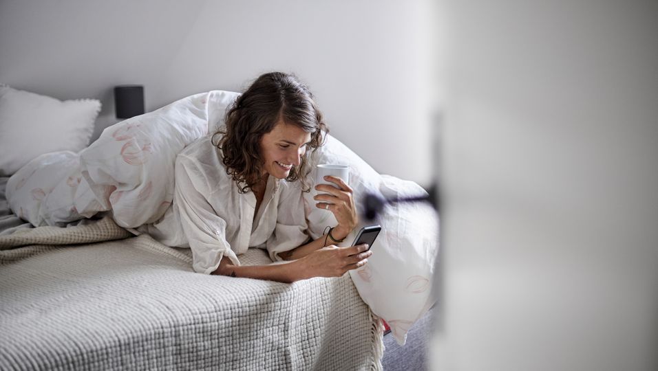 Frau liegt mit Straßenkleidung im Bett, schaut auf ihr Handy und trinkt Kaffee