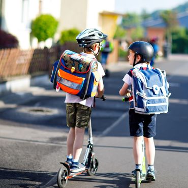 Zwei Jungs mit Schulranzen fahren mit dem Roller in die Schule.